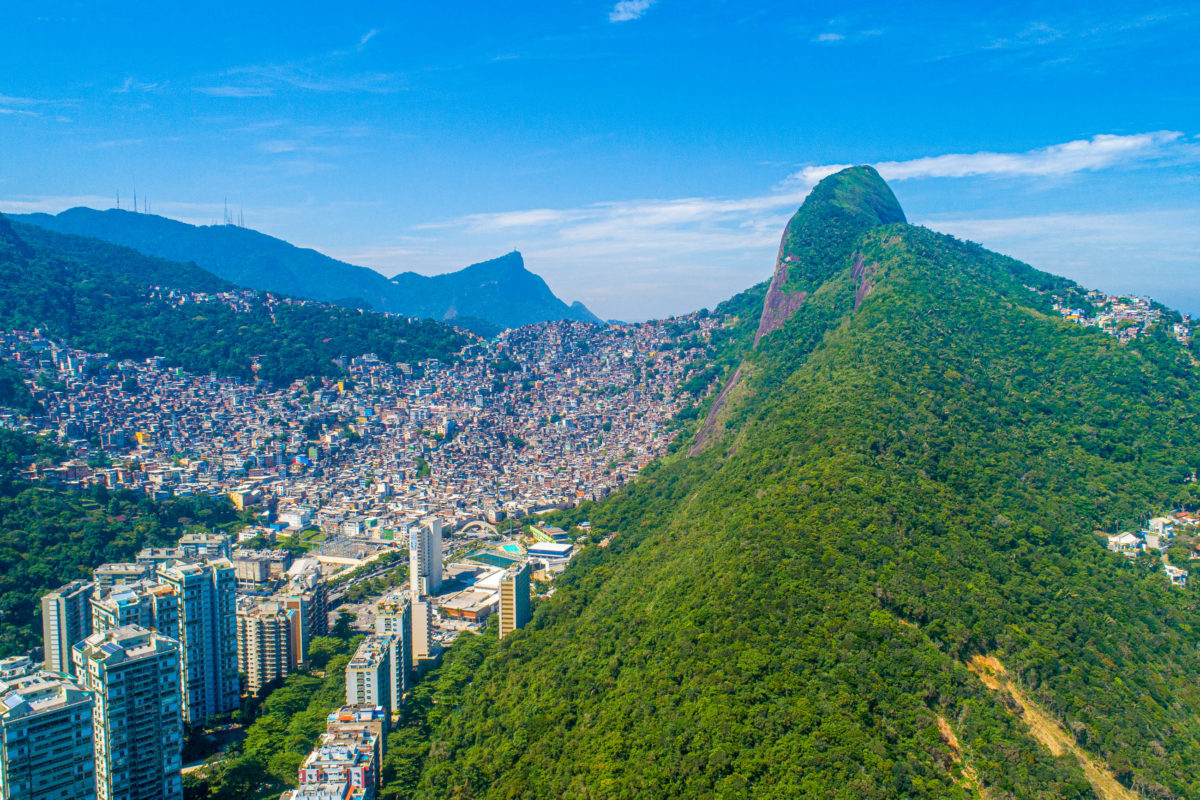 Luftaufnahme der Favela da Rocinha, dem größten Slum Brasiliens auf einem Berg in Rio de Janeiro, im Vordergrund die Skyline der Stadt - © Brastock / Shutterstock