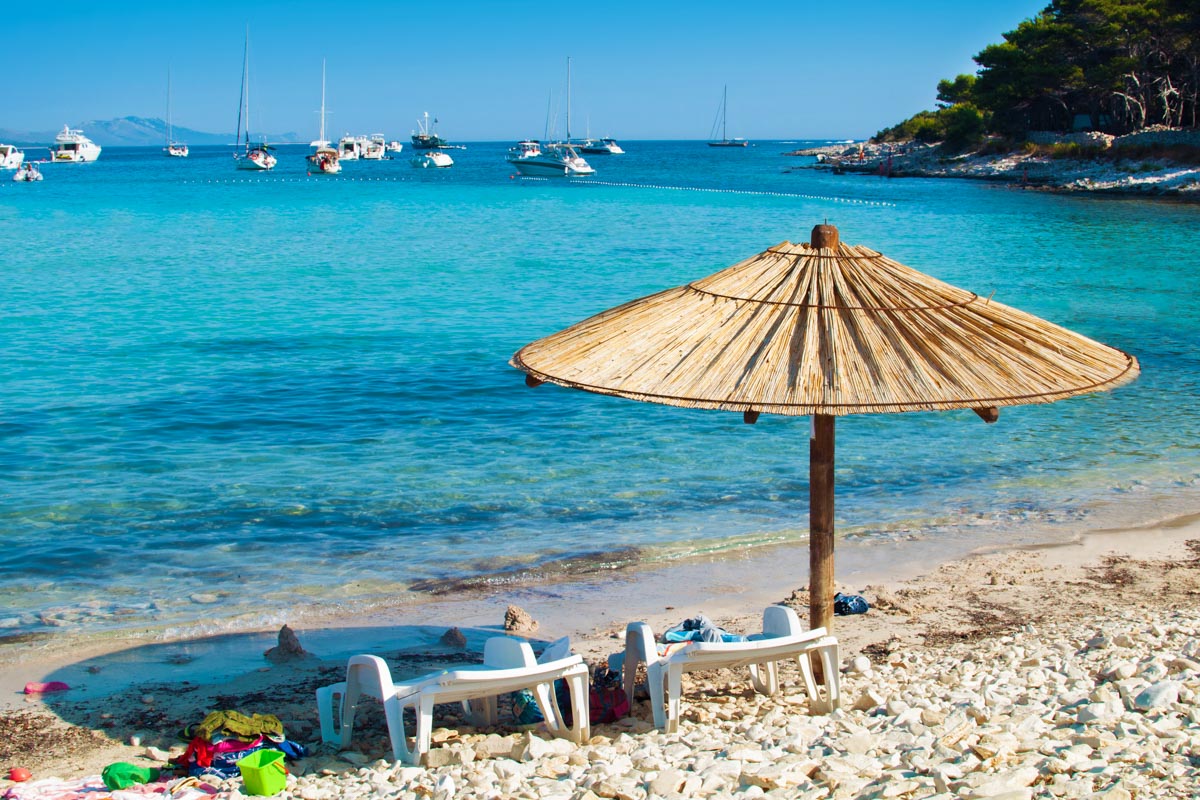 Sonnenschirm und Liegestühle am Kieselsandstrand von Dugi Otok. In der azurblauen Meereslagune ankern einige Yachten, Kroatien