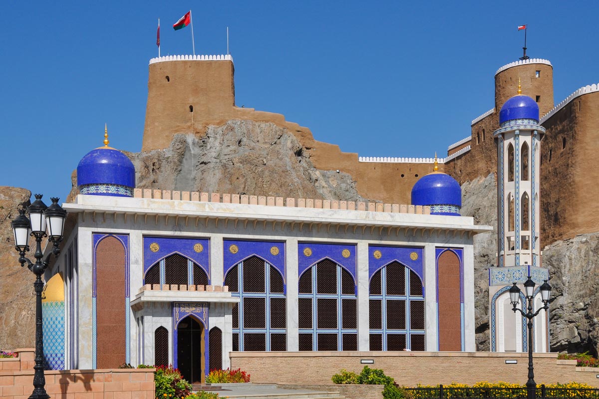 Blaue Kuppeln und blaue Kacheln dominieren die Fassade der Al Khor Moschee von Muscat, die in starkem Kontrast zum graubraunen Fort Mirani dahinter stehen, Oman